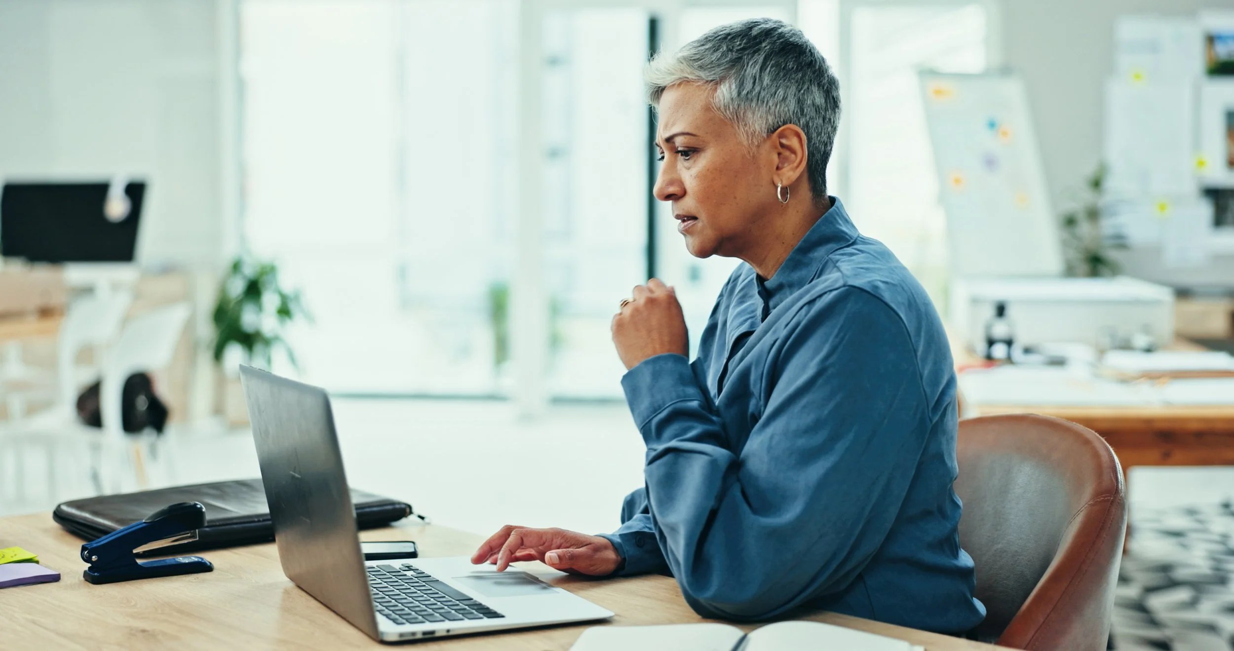 Older woman with short gray hair working on a laptop at a desk in a bright office space trying to figure out the complexity of our healthcare system.