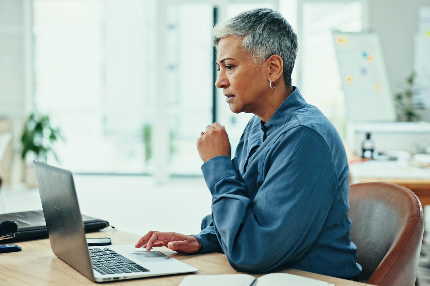 Older woman with short gray hair working on a laptop at a wooden table in a bright, modern office.
