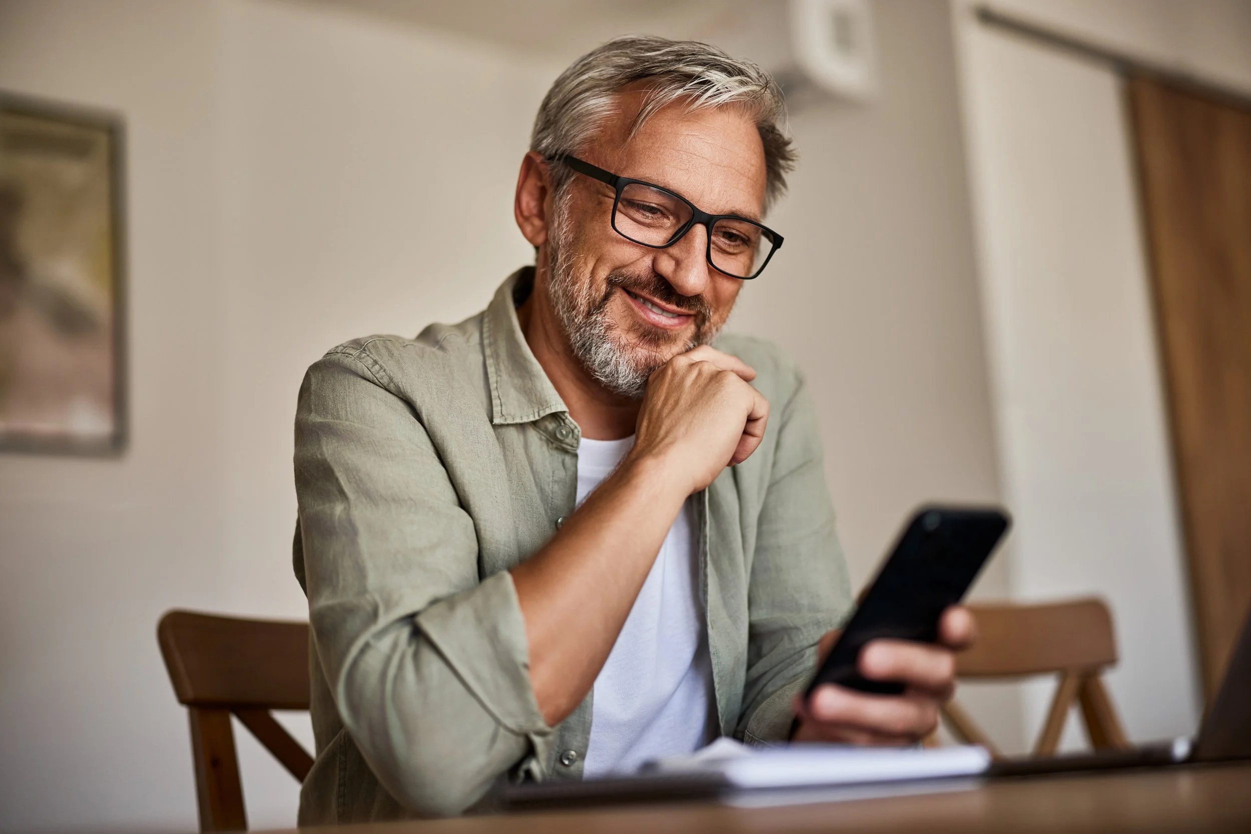An older man with gray hair and a beard, wearing glasses and a light green shirt, sitting at a wooden table, looking at his smartphone, realizing an insight about his health.