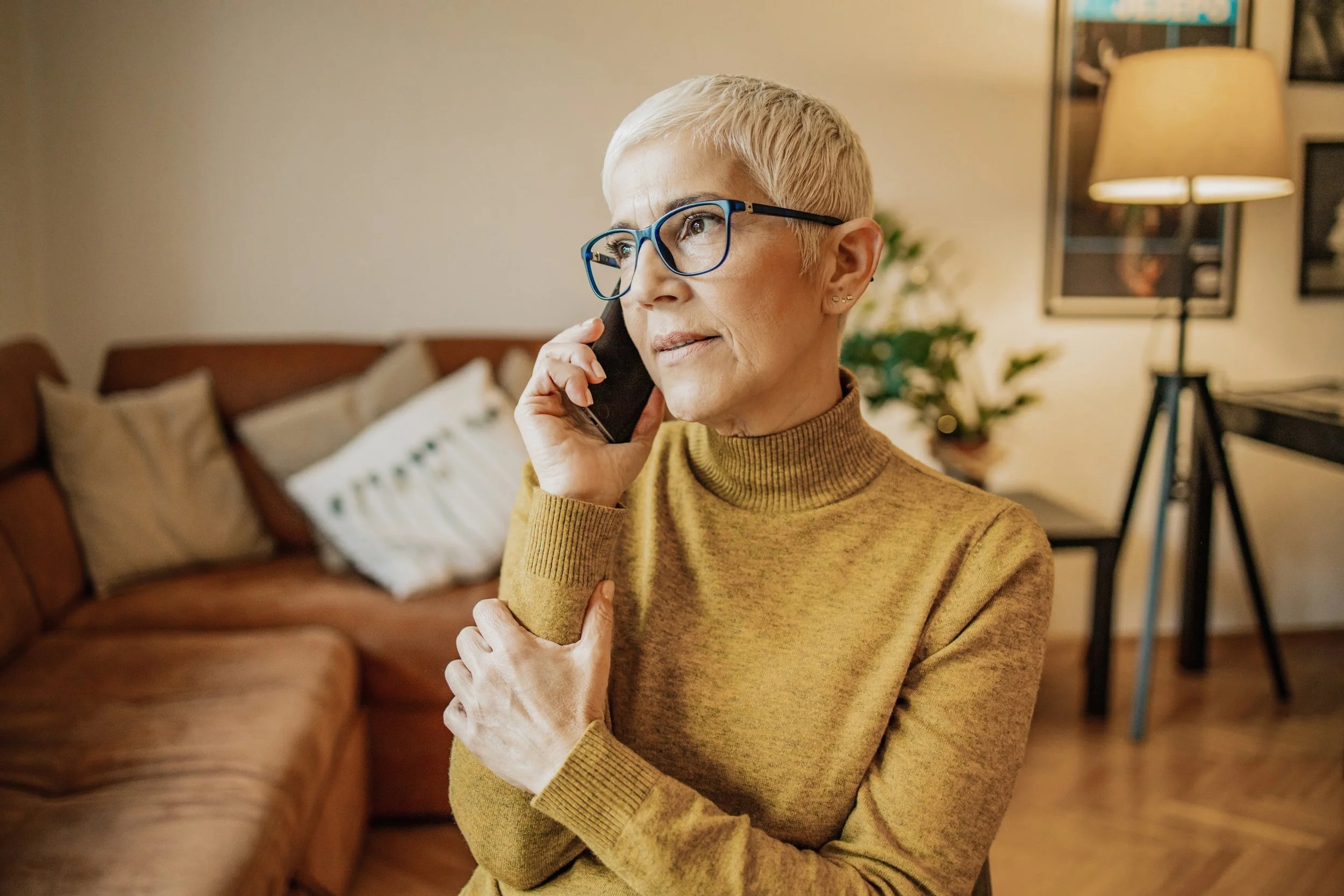 An older woman with short gray hair, glasses, and a mustard yellow turtleneck sweater talking on a cellphone asking her friends about her osteoporosis drug options.